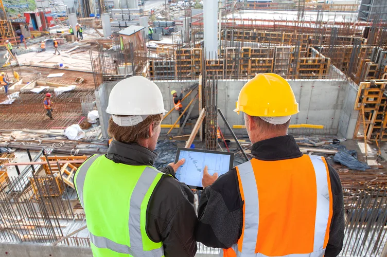 Due tecnici in casco e gilet ad alta visibilità consultano il tablet sopra un cantiere con armature in ferro